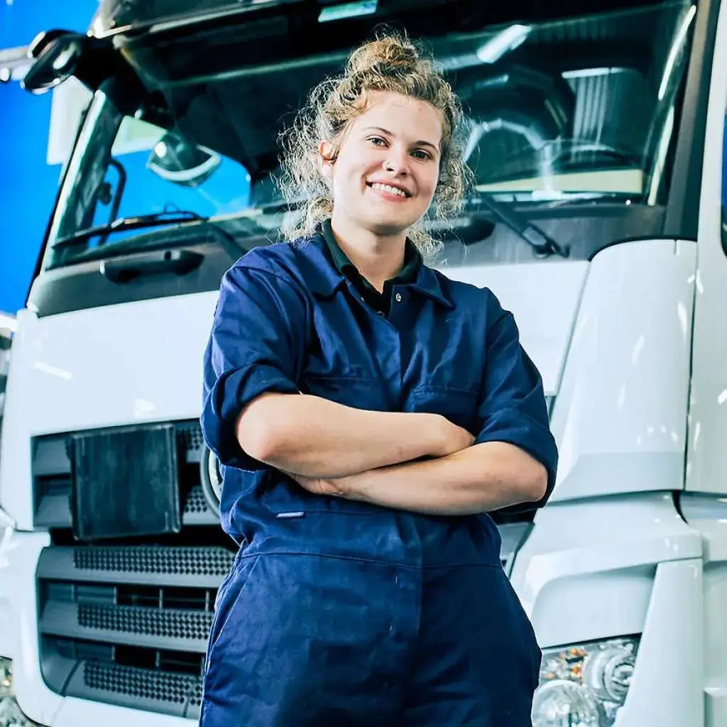 A woman in blue overalls stands confidently in front of a white lorry, smiling and crossing her arms. Education in vocational skills highlighted.
