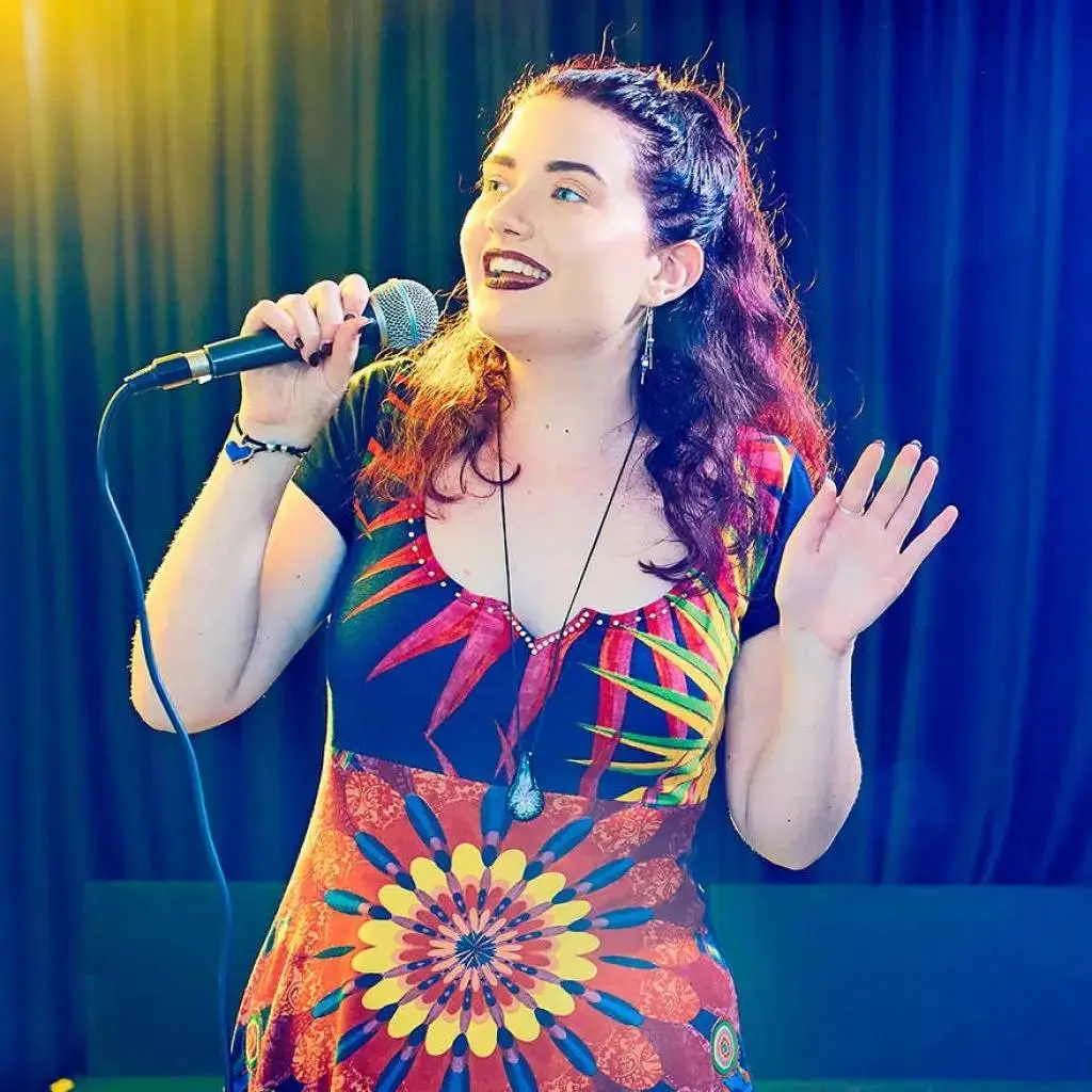 A woman in a colourful dress singing into a microphone on stage with a blue curtain backdrop.