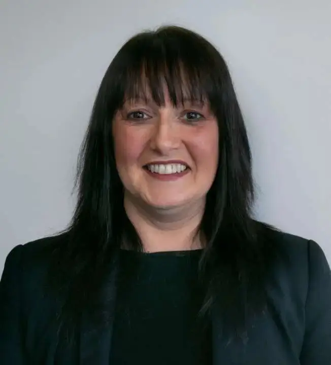 A woman with long dark hair and bangs smiles at the camera, wearing a black blazer over a black top. She stands against a plain white background.