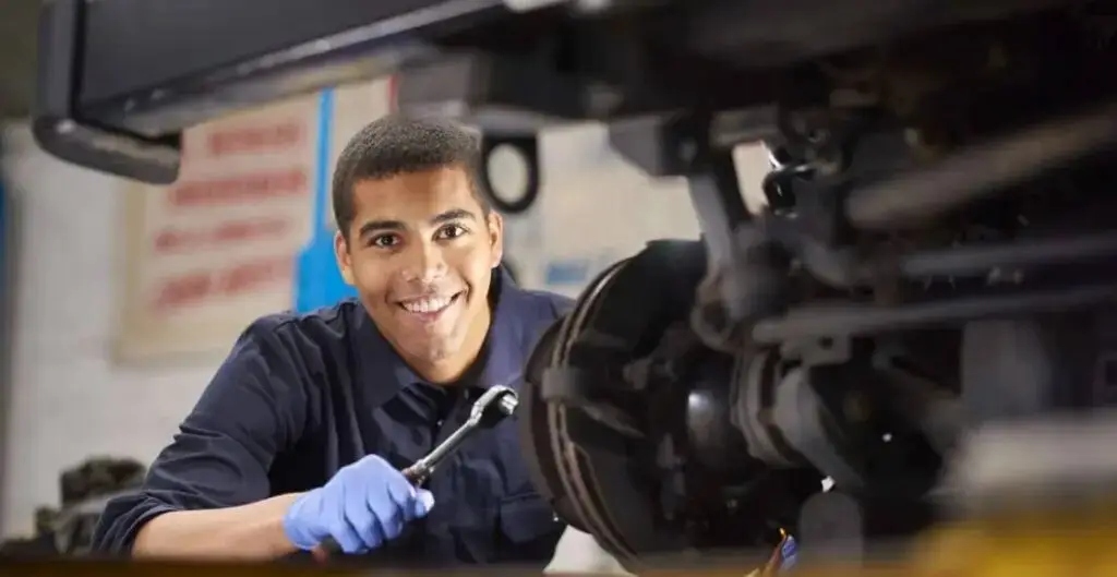 A student in a blue uniform and gloves works on a car's brake system in a well-lit garage, smiling at the camera.