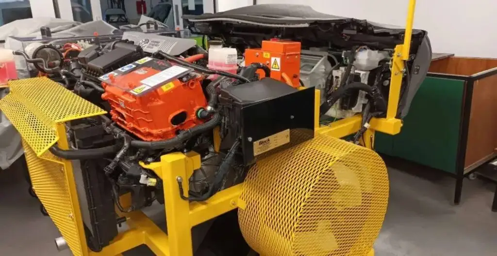 A yellow engine stand holds a red engine with black components and a yellow fan, likely used for educational purposes in a UK college automotive programme.