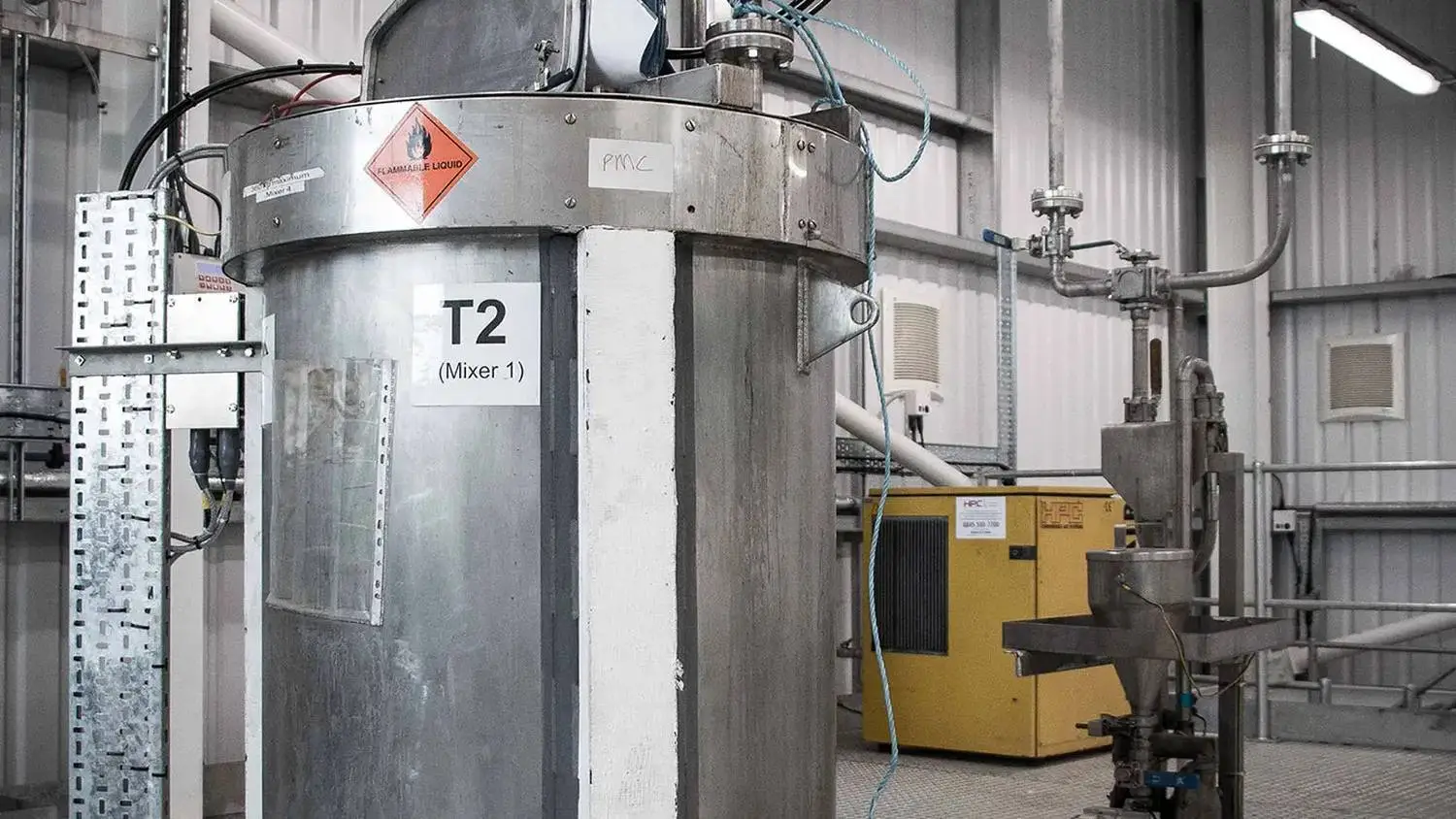 Industrial mixer labeled 'T2 (Mixer 1)' in a laboratory setting, with pipes and safety signage.