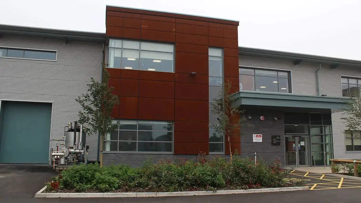 Modern college building with red and grey facade, large windows, and landscaped entrance. Signage and accessible parking visible.