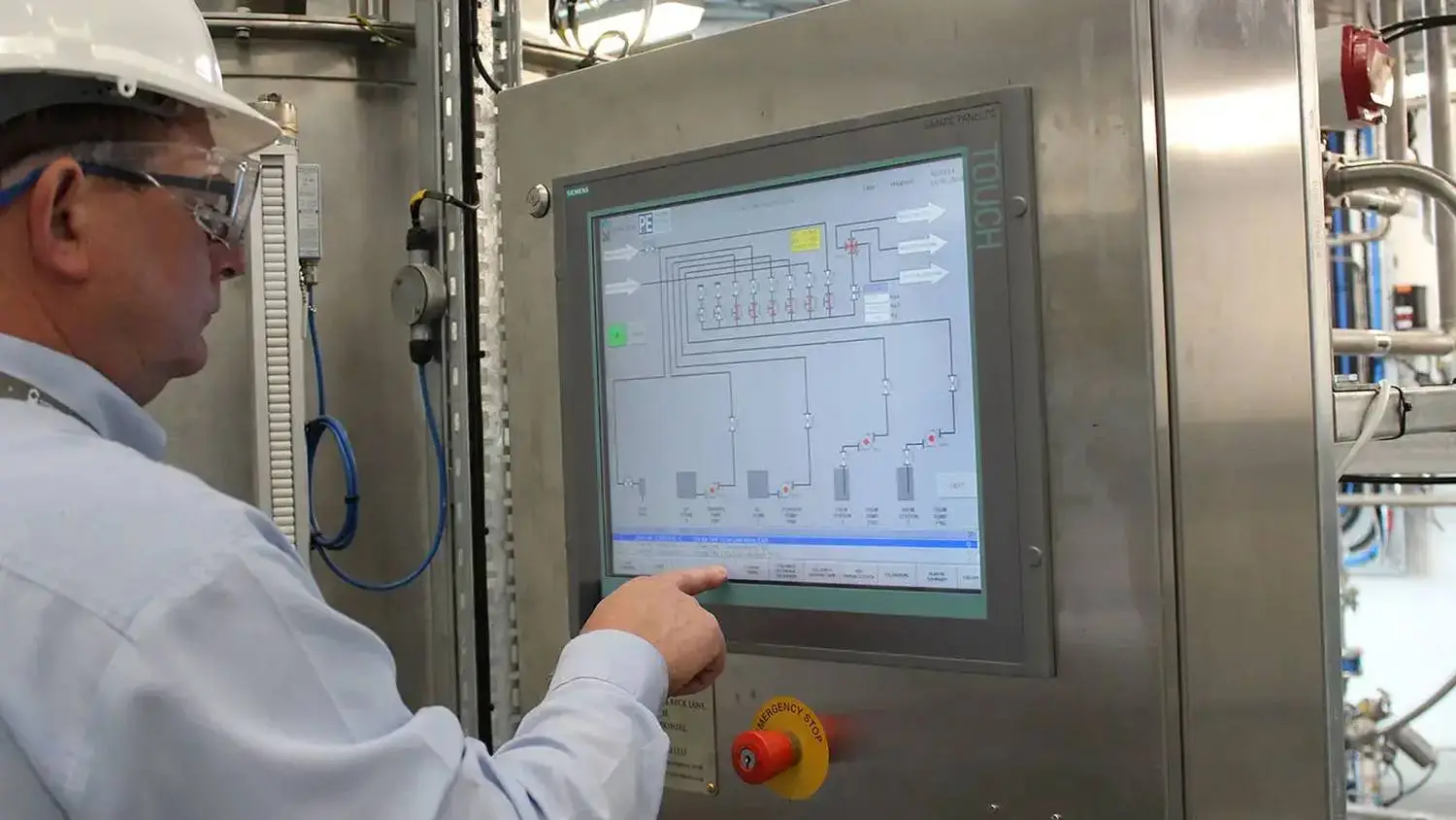Person in hard hat and safety glasses operating industrial control panel with schematic diagram.