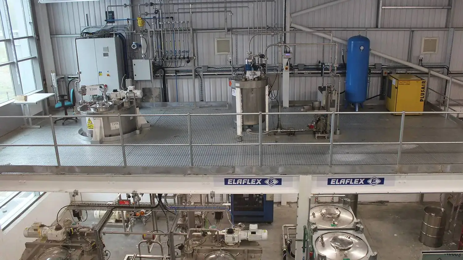 Industrial processing facility with stainless steel equipment, pipes, and control panels. Elevated walkway and blue storage tank visible. ELAFLEX signage prominent.