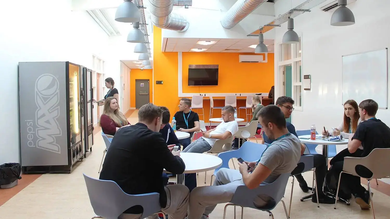 Students in a modern college break room, seated around tables with laptops and notebooks, engaged in discussion under industrial-style lighting.