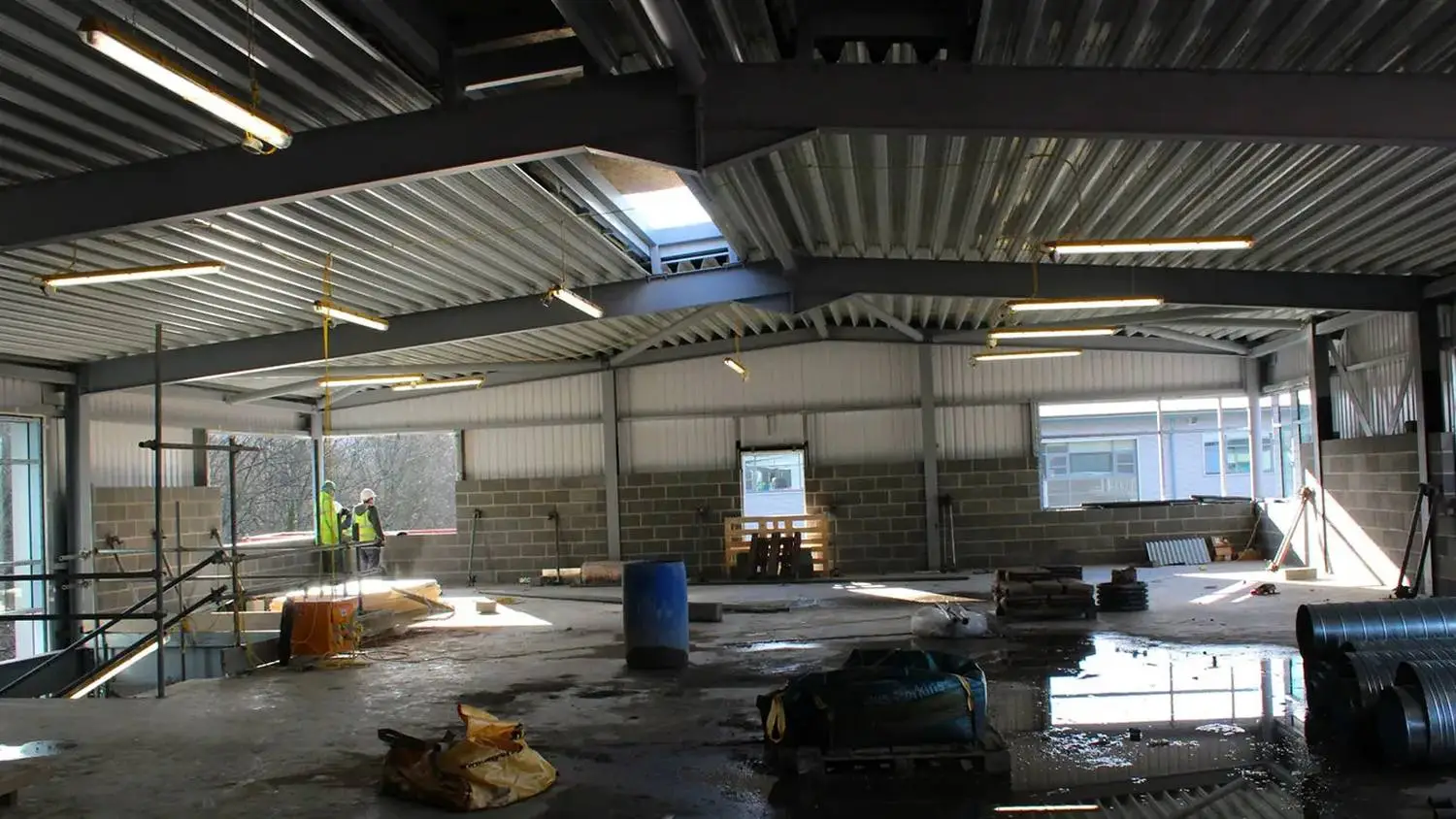 Construction site inside a large industrial building with exposed brick walls, metal beams, and workers in high-visibility vests. Puddles and building materials scattered on the concrete floor.