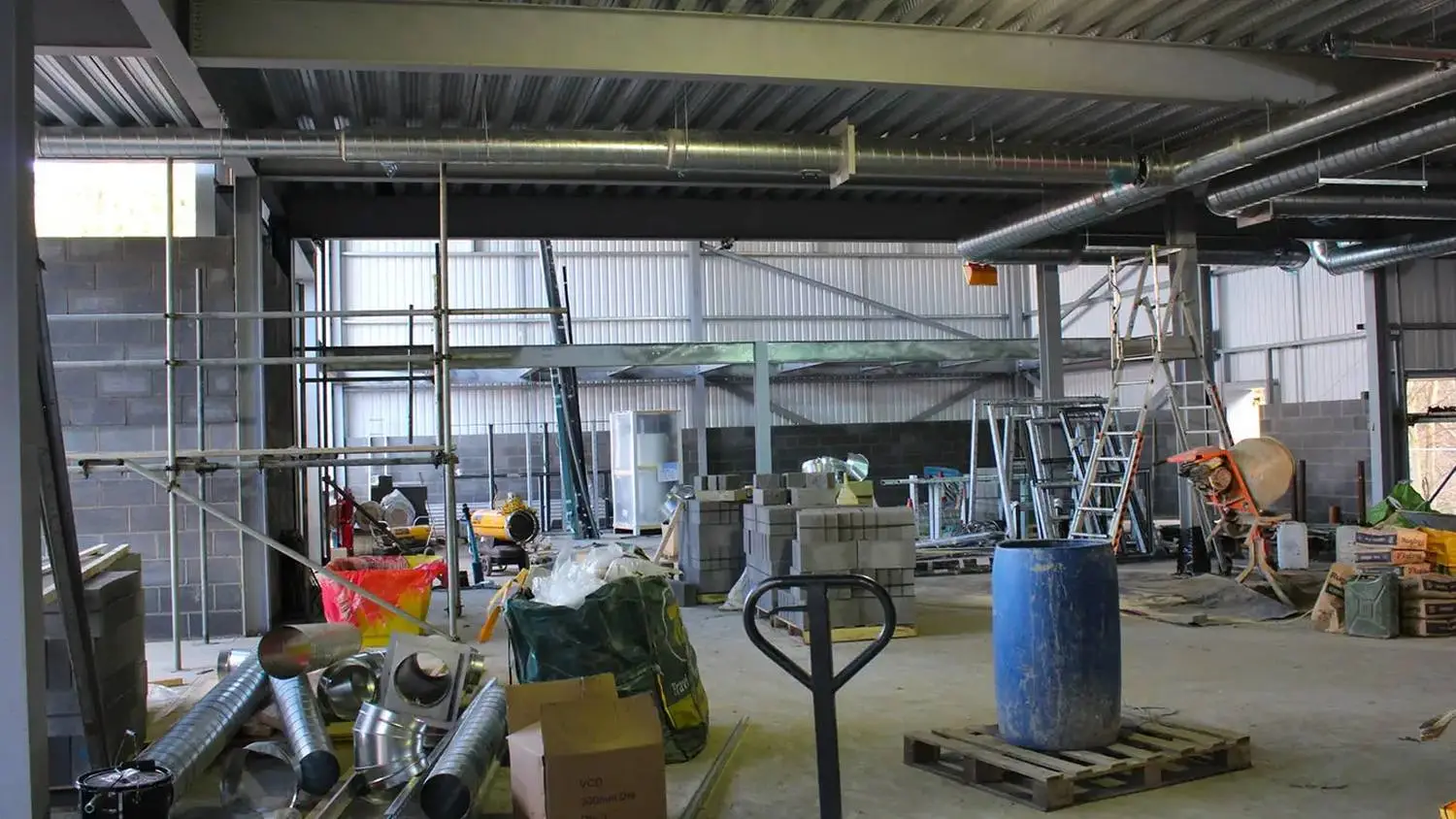 A college construction site with scaffolding, ladders, ductwork, and building materials under a metal roof.