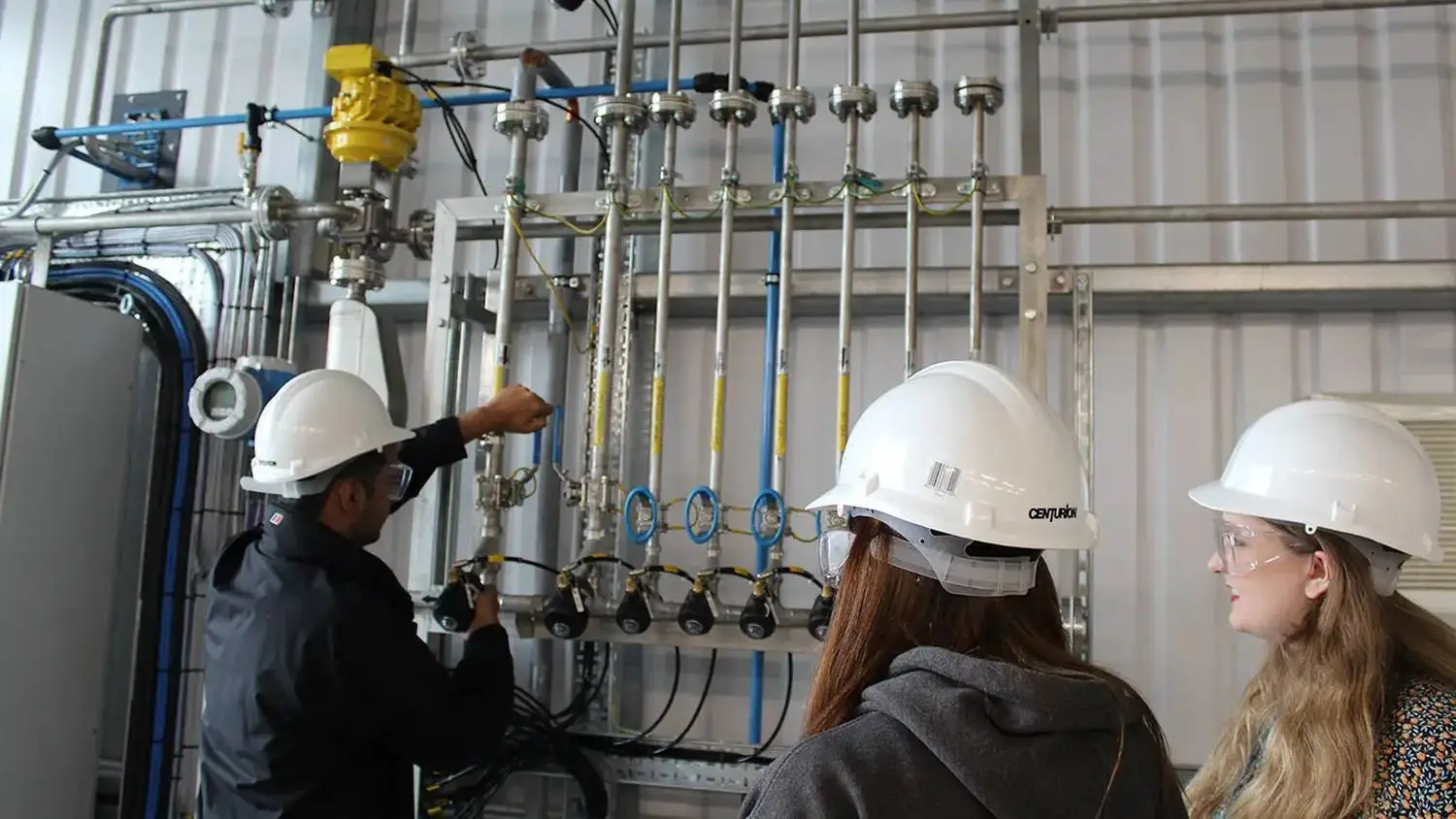 A man in a hard hat explaining a complex pipe system to two students in hard hats and safety glasses.