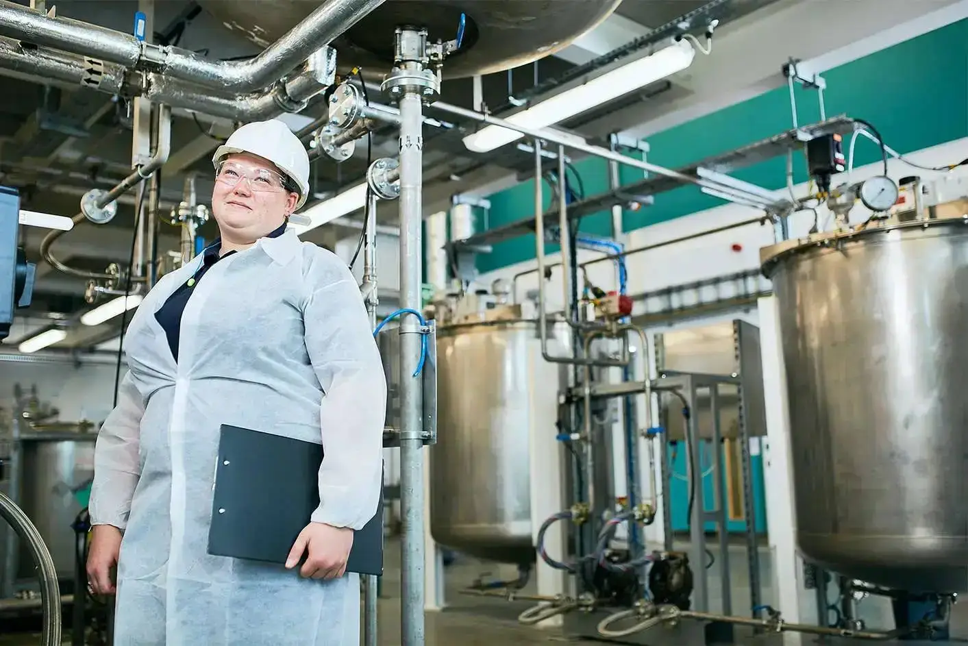 A person in a white lab coat and hard hat holding a folder stands in front of industrial tanks and pipes in a well-lit facility.
