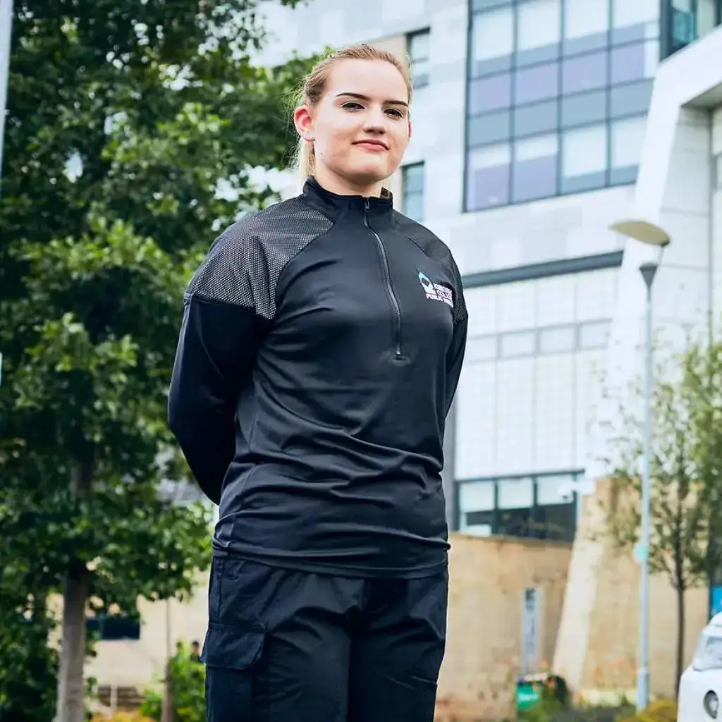 A person in black sportswear stands confidently in front of a modern college building with trees and a clear sky.