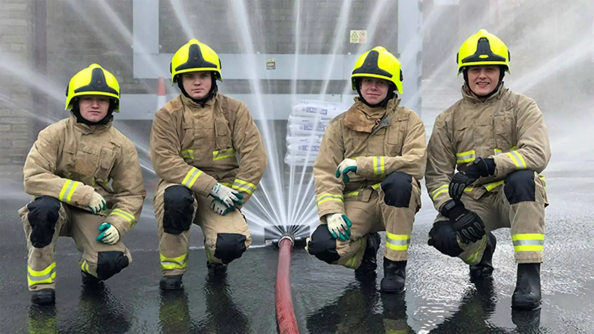 Four firefighters in protective gear kneeling beside a hose spraying water, demonstrating firefighting techniques in a training exercise.
