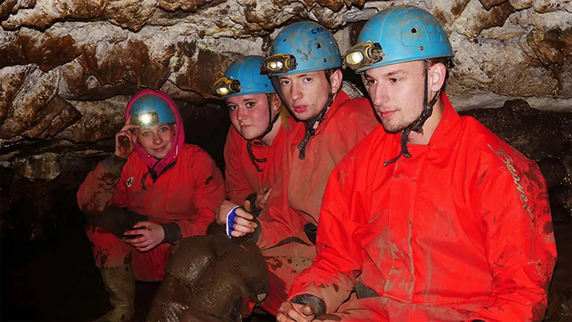 Four people in red overalls and blue helmets with headlamps crouch in a rocky cave, engaged in a caving expedition.