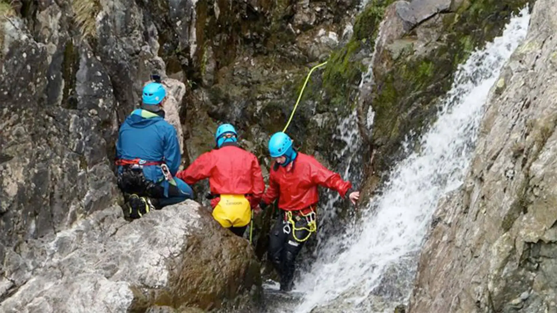 Three people in helmets and harnesses climbing a waterfall, demonstrating outdoor adventure education techniques in a rugged, natural setting.