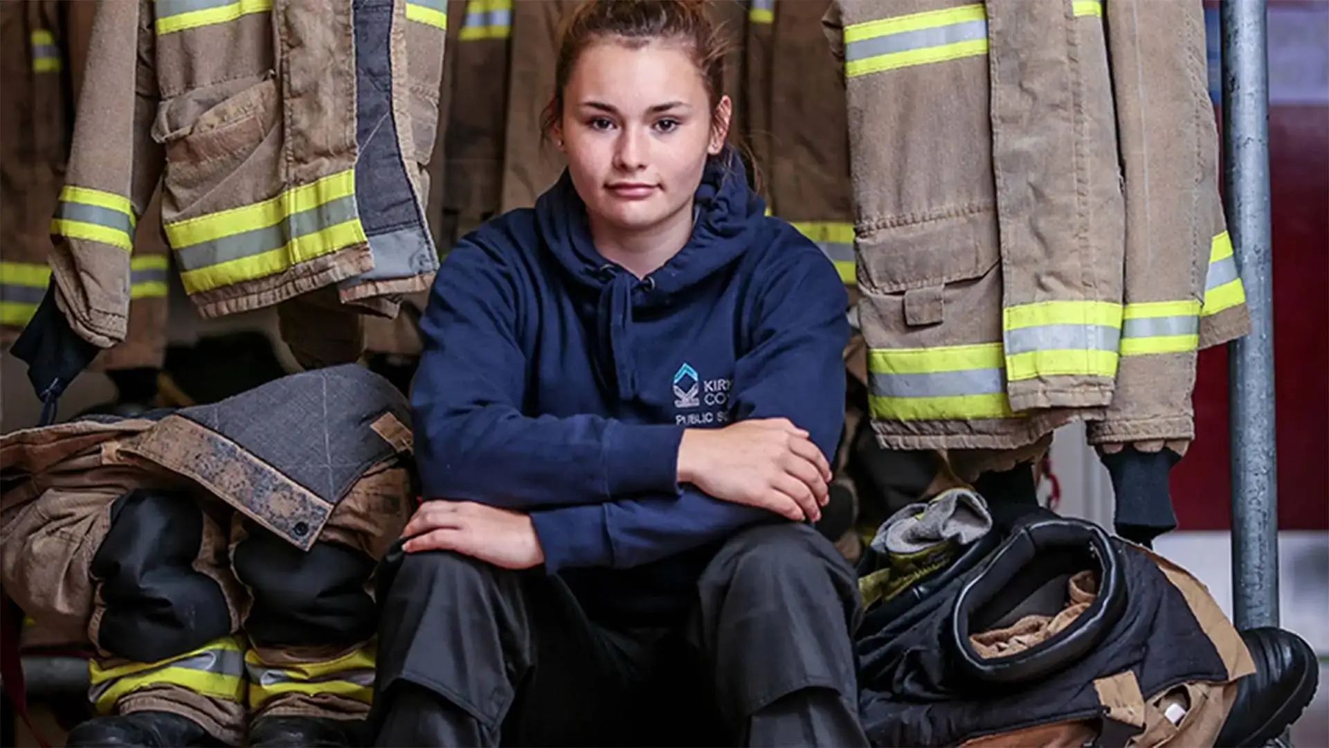A young woman in a blue hoodie sits in front of firefighter uniforms, symbolising career aspirations and educational pathways in emergency services.