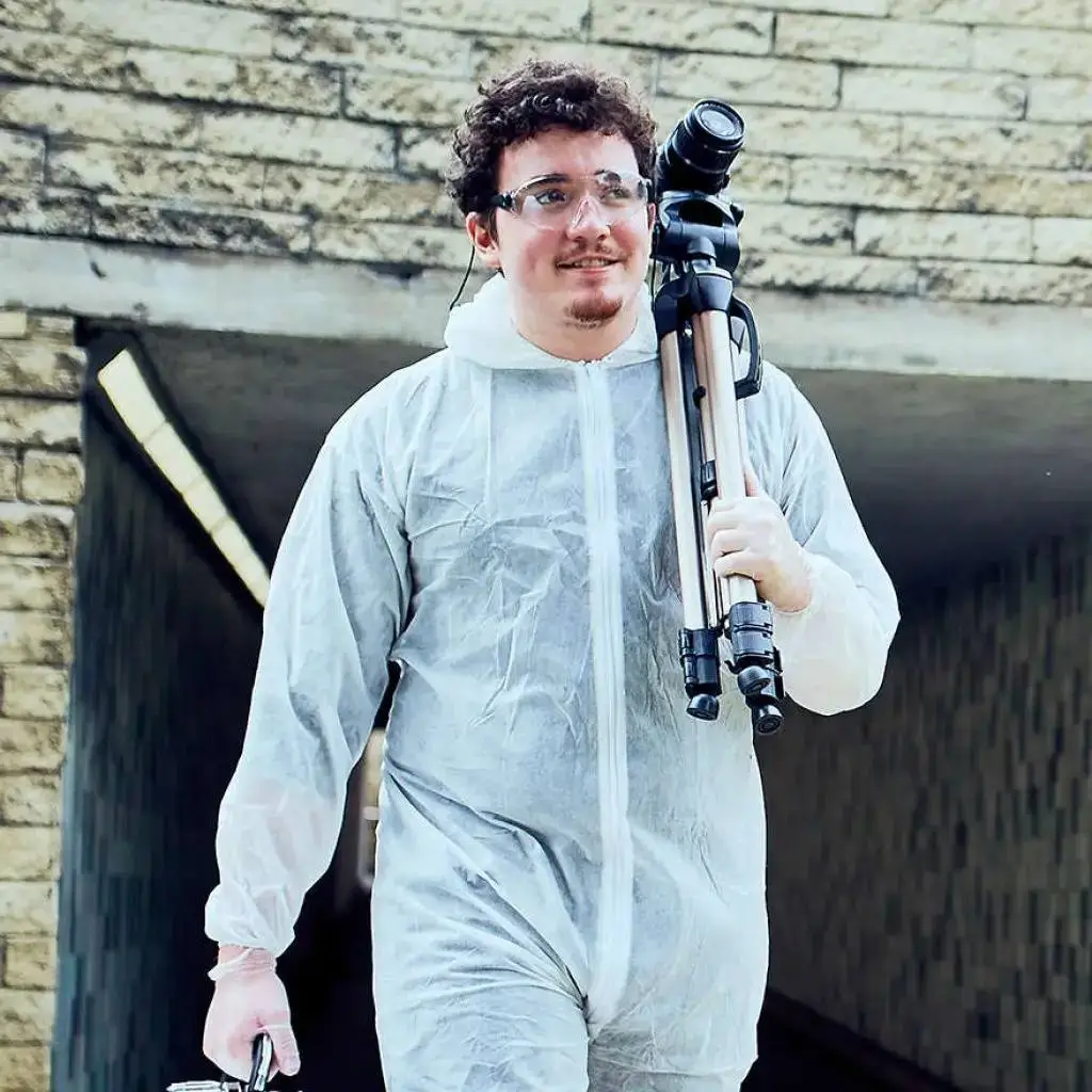 A person in a white protective suit holding a camera on a tripod, standing in front of a stone building.