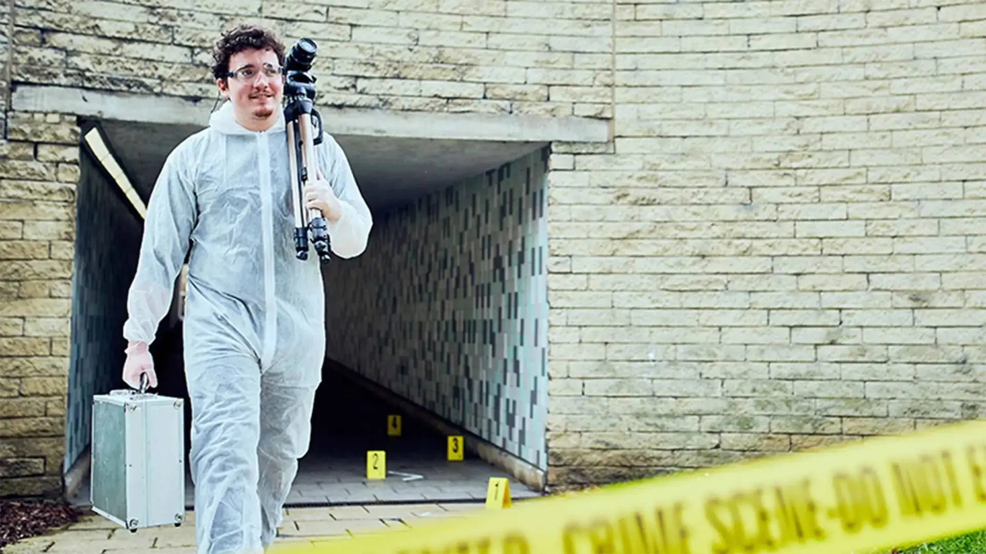 A person in a white protective suit carrying a camera and a case exits a brick building entrance marked with crime scene tape.