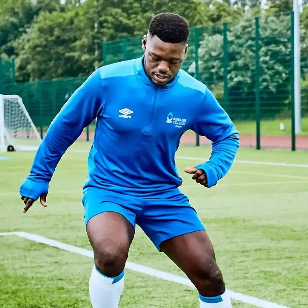 A person in a blue sports outfit practising football on a green pitch, with a goalpost and trees in the background.