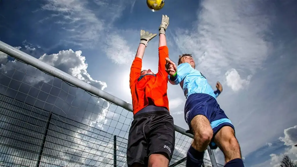 Two football players in a match, one in orange and one in blue, jumping to head a yellow ball near a goalpost under a blue sky with clouds.