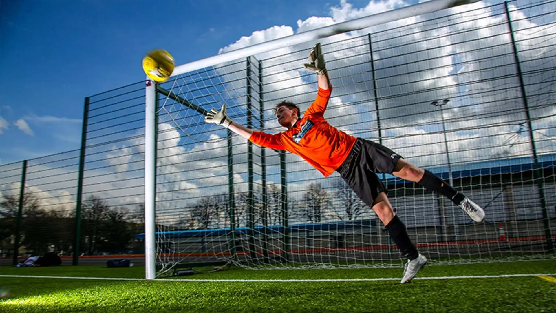 A goalkeeper in an orange jersey dives to save a yellow football against a goalpost on a sunny day.
