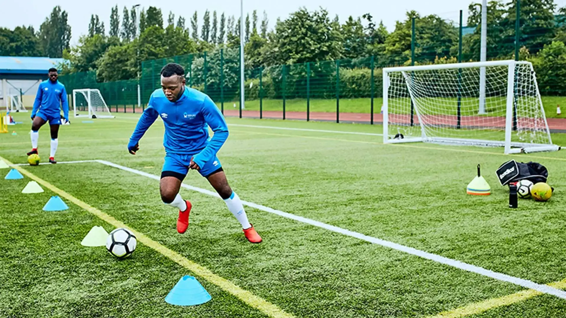 Two football players in blue training kits practising dribbling through cones on a green pitch near a goalpost.