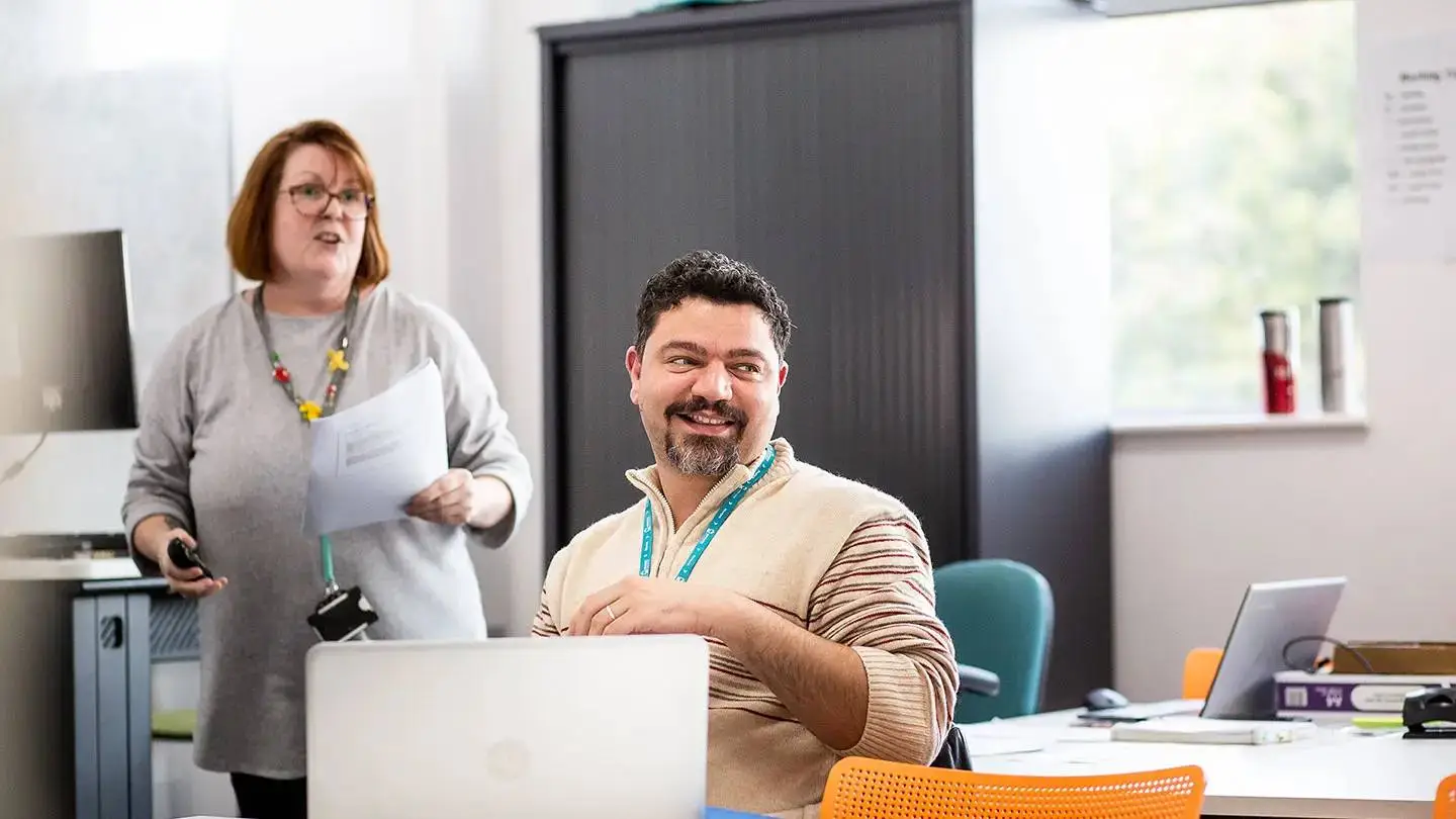 Two people in a college classroom, one seated with a laptop, the other standing holding papers, both smiling.