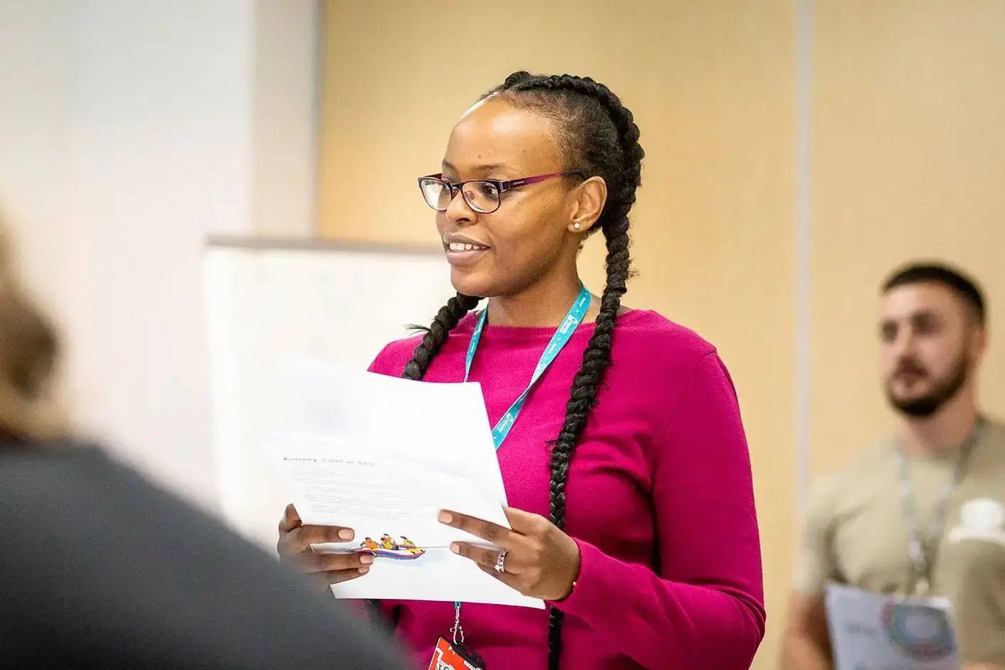 A woman in a pink jumper holding papers, smiling and speaking to a group in a classroom setting.