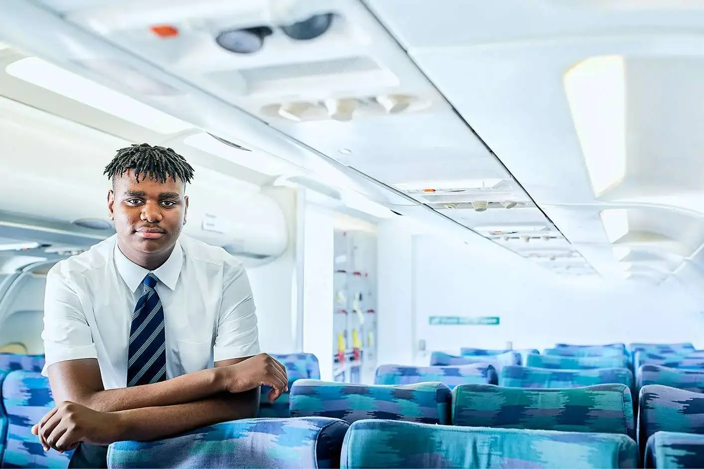 A young man in a white shirt and tie sits in an empty aeroplane cabin, looking towards the camera.