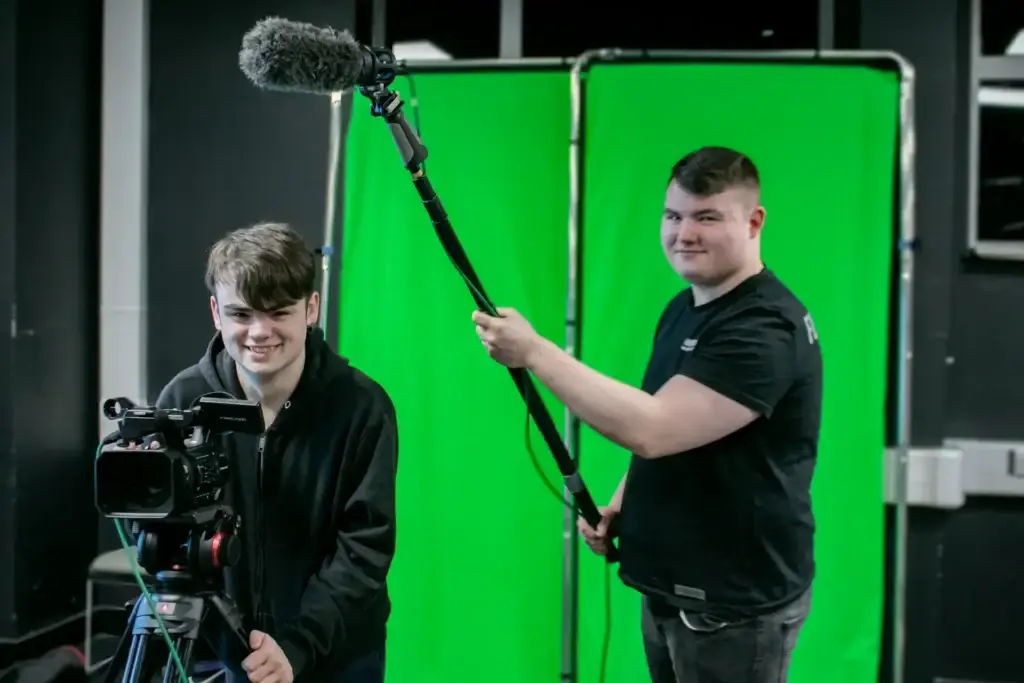 Young man operates camera and another operates boom mic to the left in front of green screen