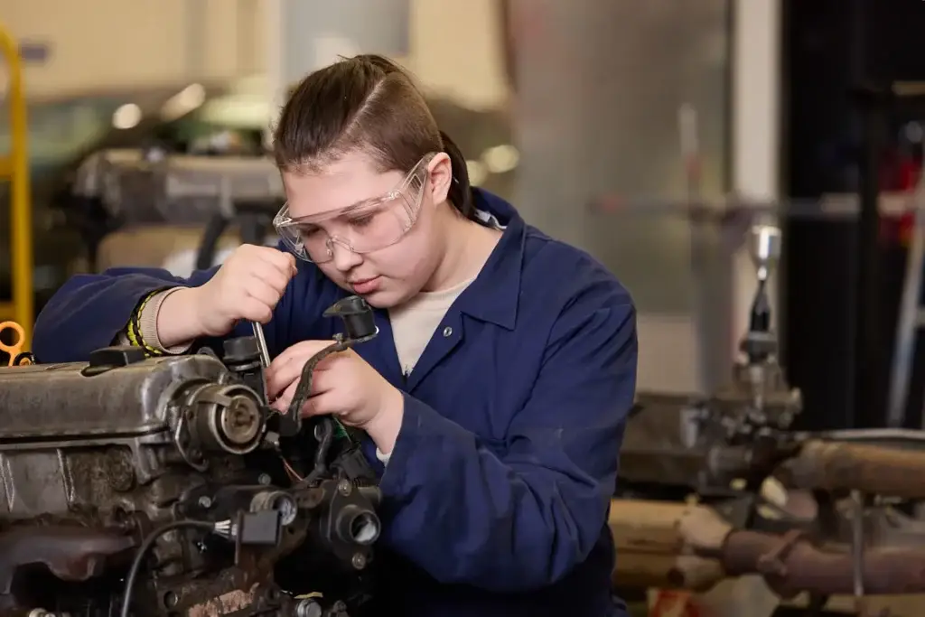 A student wearing goggles and blue overalls working on piece of machinery.