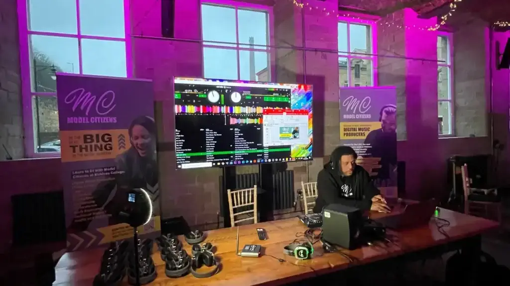 Man DJing at wooden table in front of plasma screen showing DJ mixing