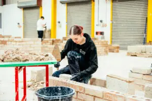 Student kneels next to brick wall holding tool