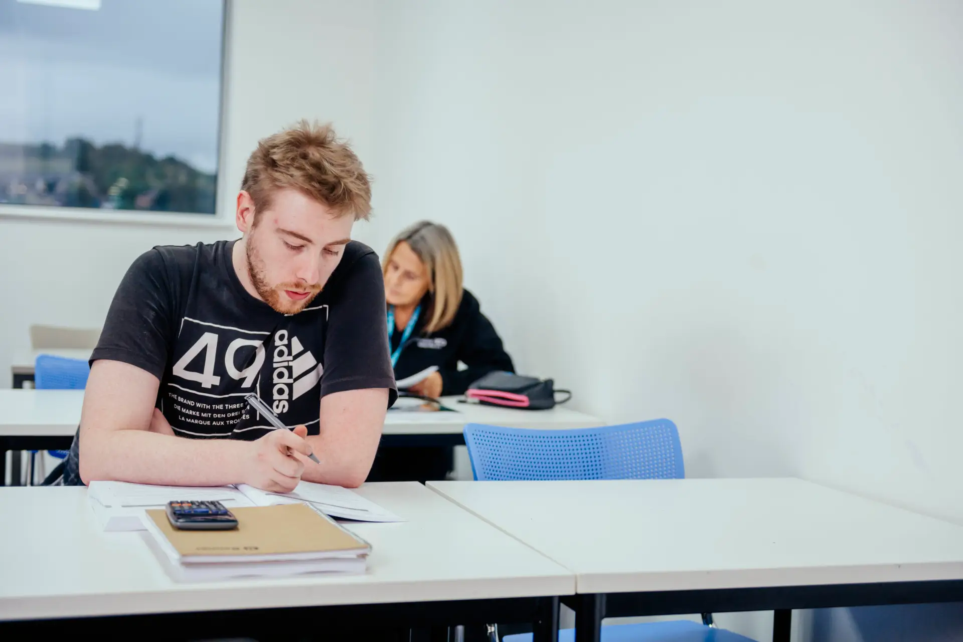 Man sits at a desk writing in a notebook with woman in the background