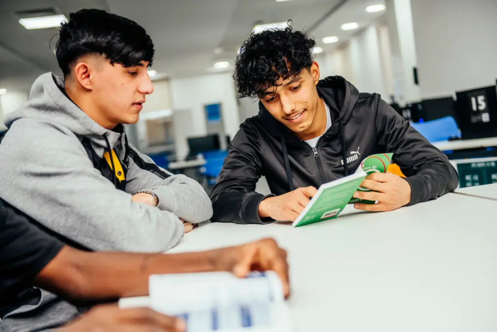 Twp young men sit at a desk reading a green book