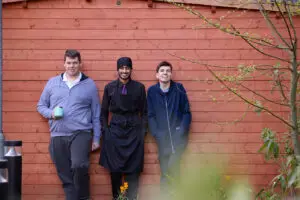 3 students leant against wooden wall