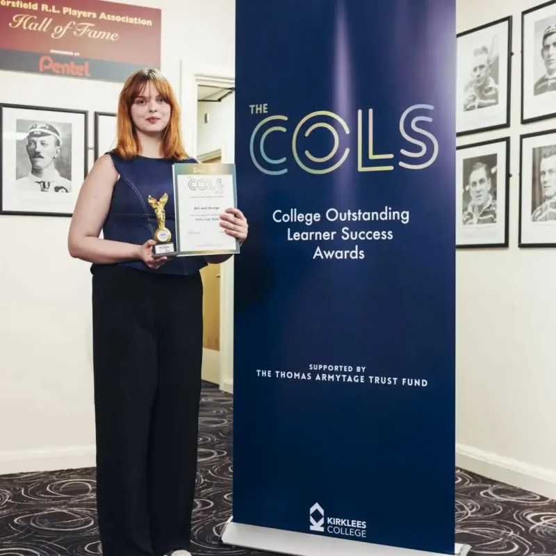 A student in a dark outfit holds a trophy and certificate in front of a COLS awards banner at Kirklees College.