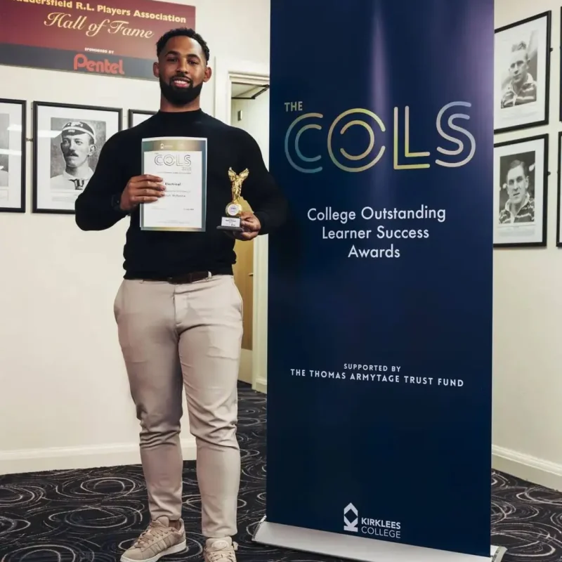 A man in a black sweater holding a trophy and certificate stands next to a blue banner for the College Outstanding Learner Success Awards.