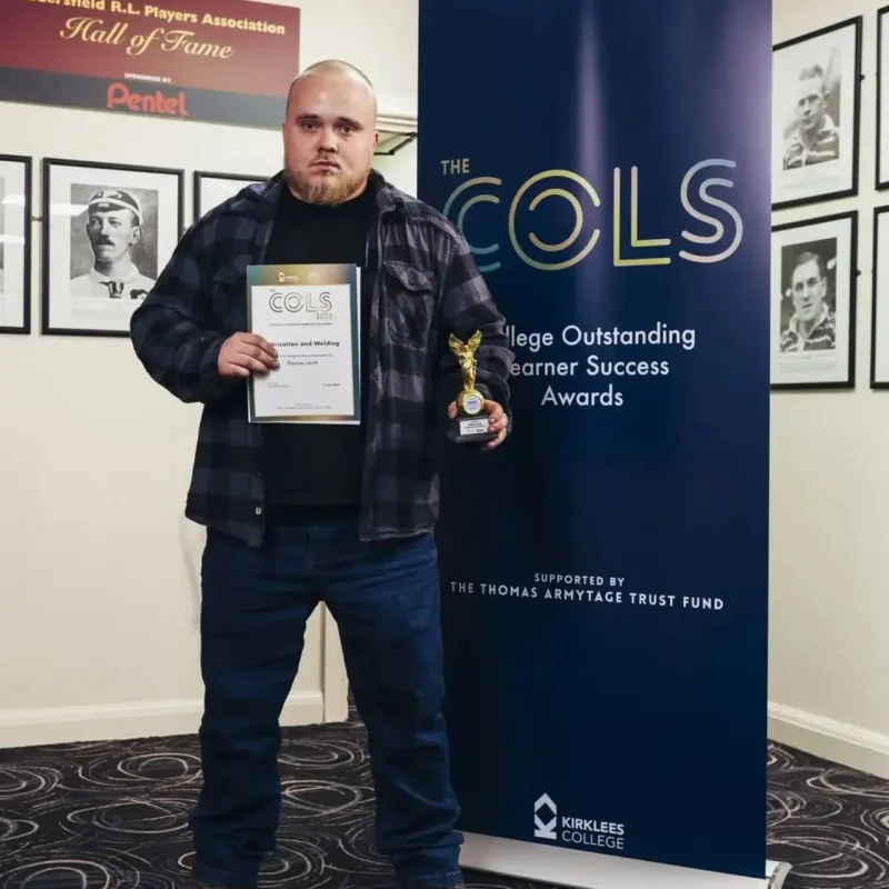 A man in a plaid shirt holding a certificate and trophy in front of a COLS banner at Kirklees College.