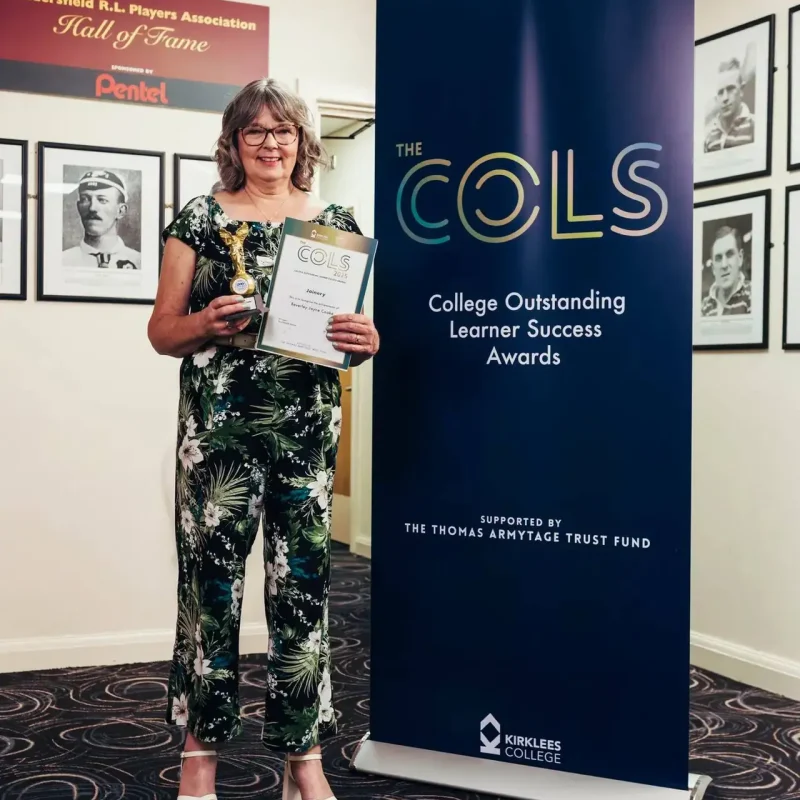A woman in a floral jumpsuit holds a certificate in front of a COLS banner at Kirklees College, celebrating learner success.