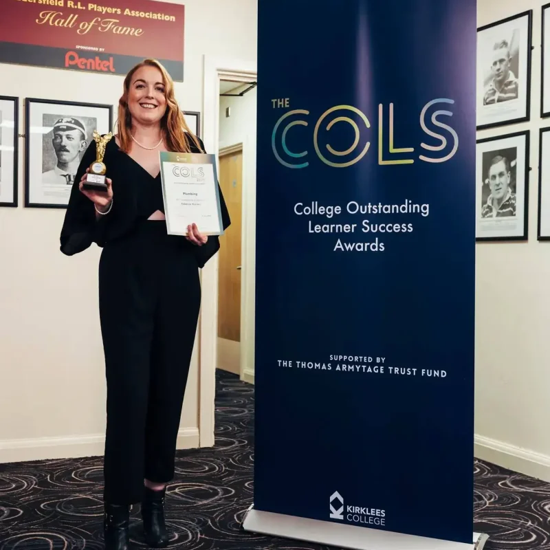 A woman in black holding a trophy and certificate stands next to a blue banner for the College Outstanding Learner Success Awards at Kirklees College.