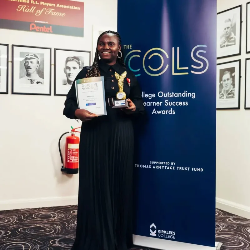 A person in a black uniform holding a certificate and trophy stands in front of a blue banner at Kirklees College, celebrating an achievement.
