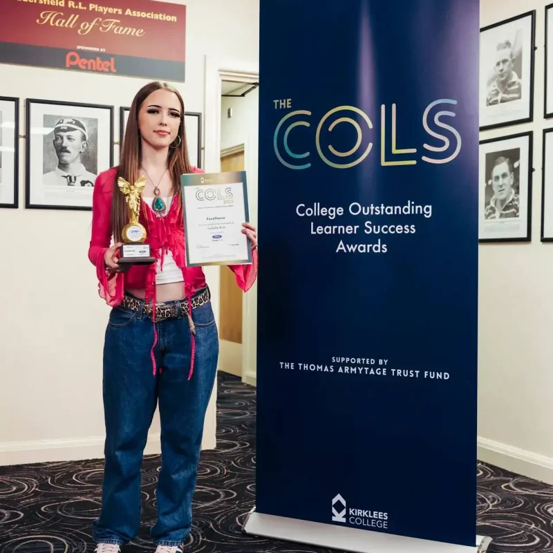 A student in a pink jacket holds a trophy and certificate in front of a COLS banner, celebrating college achievements.
