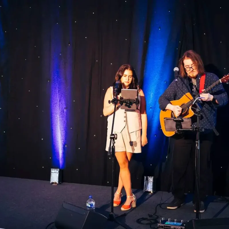 Two people on stage: one singing into a microphone with a tablet, the other playing an acoustic guitar. Blue and purple stage lighting creates a vibrant atmosphere.