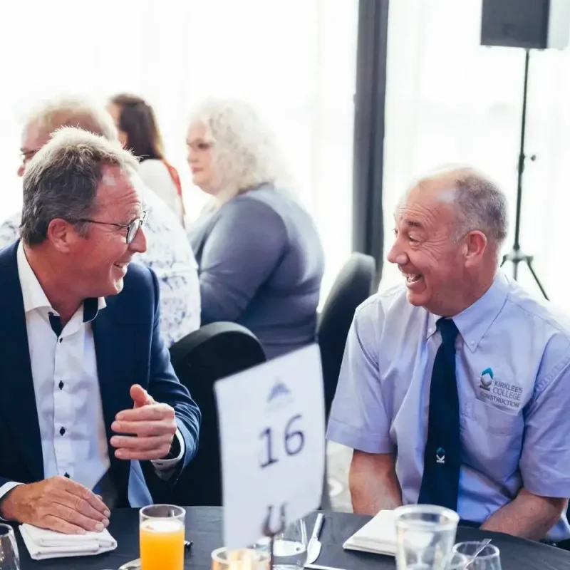 Two men in business attire smiling and talking at a table with drinks, numbered 16, in a bright room with people in the background.