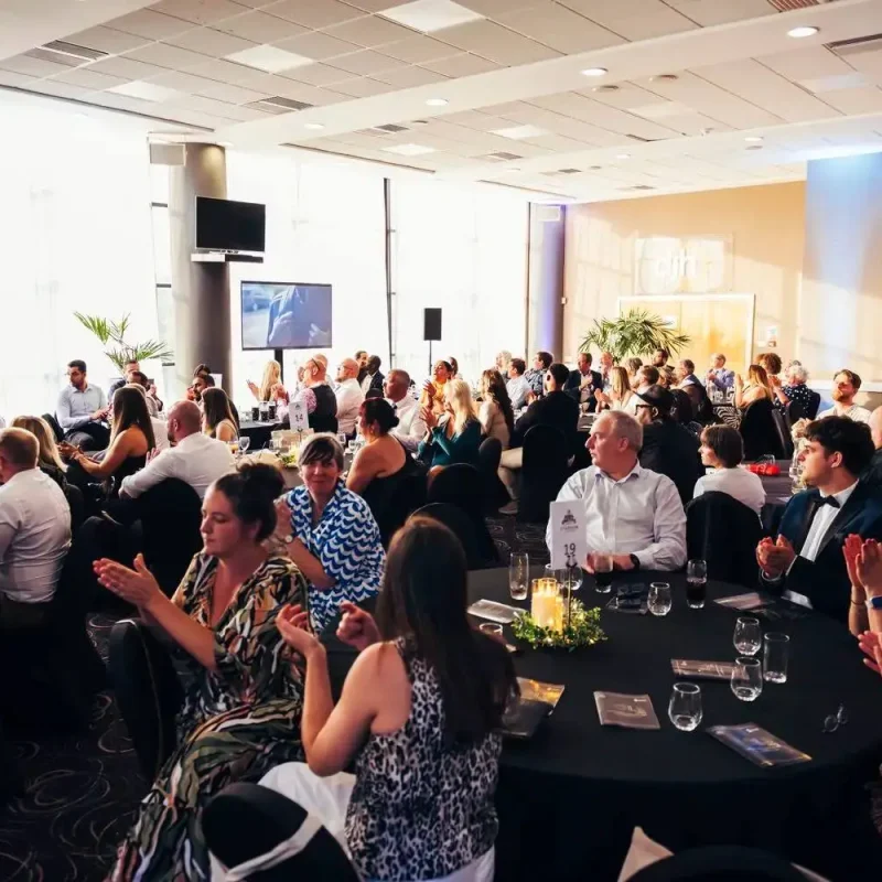 A college awards ceremony in a bright room with round tables, people clapping, and screens displaying event details.