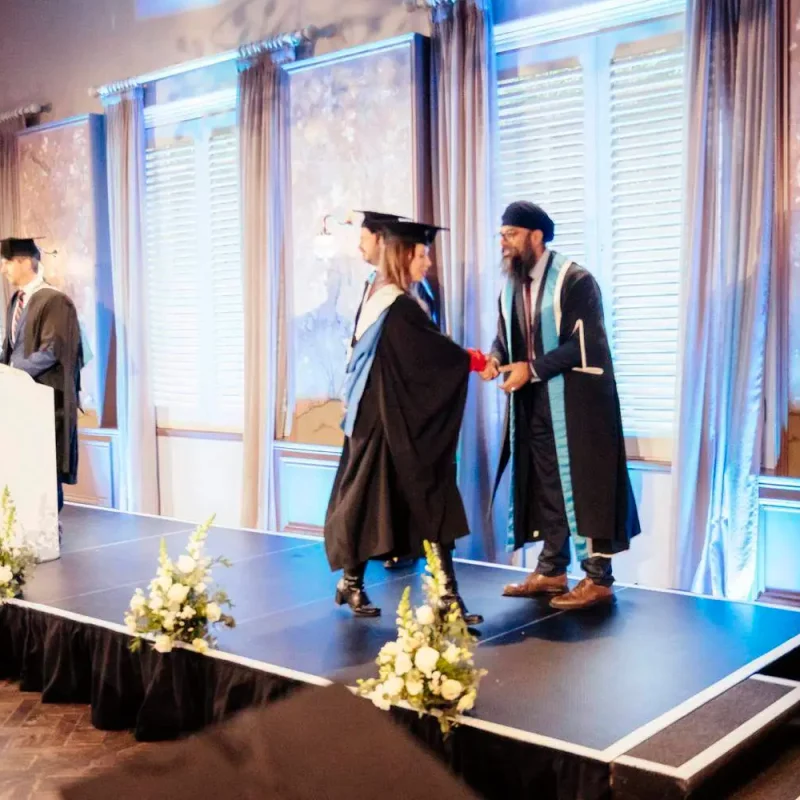 Graduates in academic gowns and caps walk across a stage, receiving certificates in a formal ceremony setting.