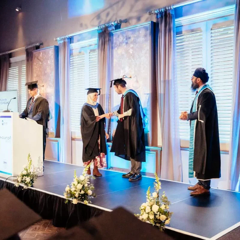 Graduates in caps and gowns receiving certificates on a stage with flowers and a podium, under blue lighting and elegant decor.