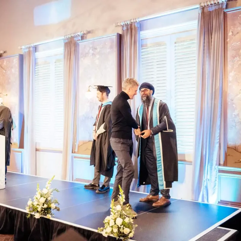 Graduates in academic gowns receiving certificates on a stage with floral arrangements and a podium.