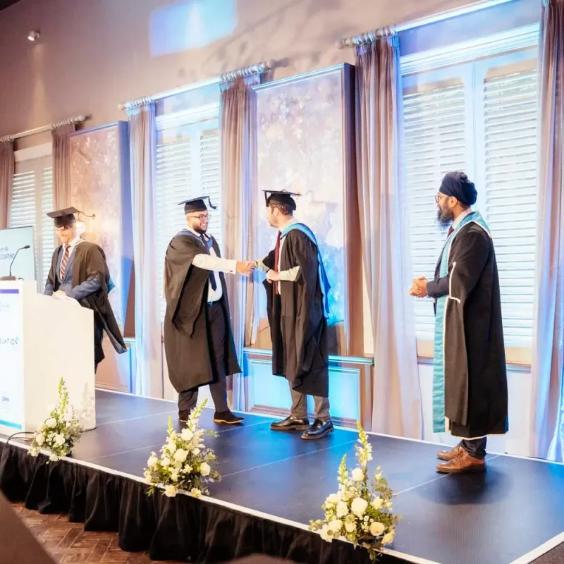 Graduates in academic gowns receiving certificates on a stage with floral arrangements and a podium, under soft lighting.