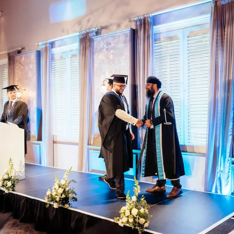 Graduation ceremony on stage with two men shaking hands, one in academic gown, podium with Graduation